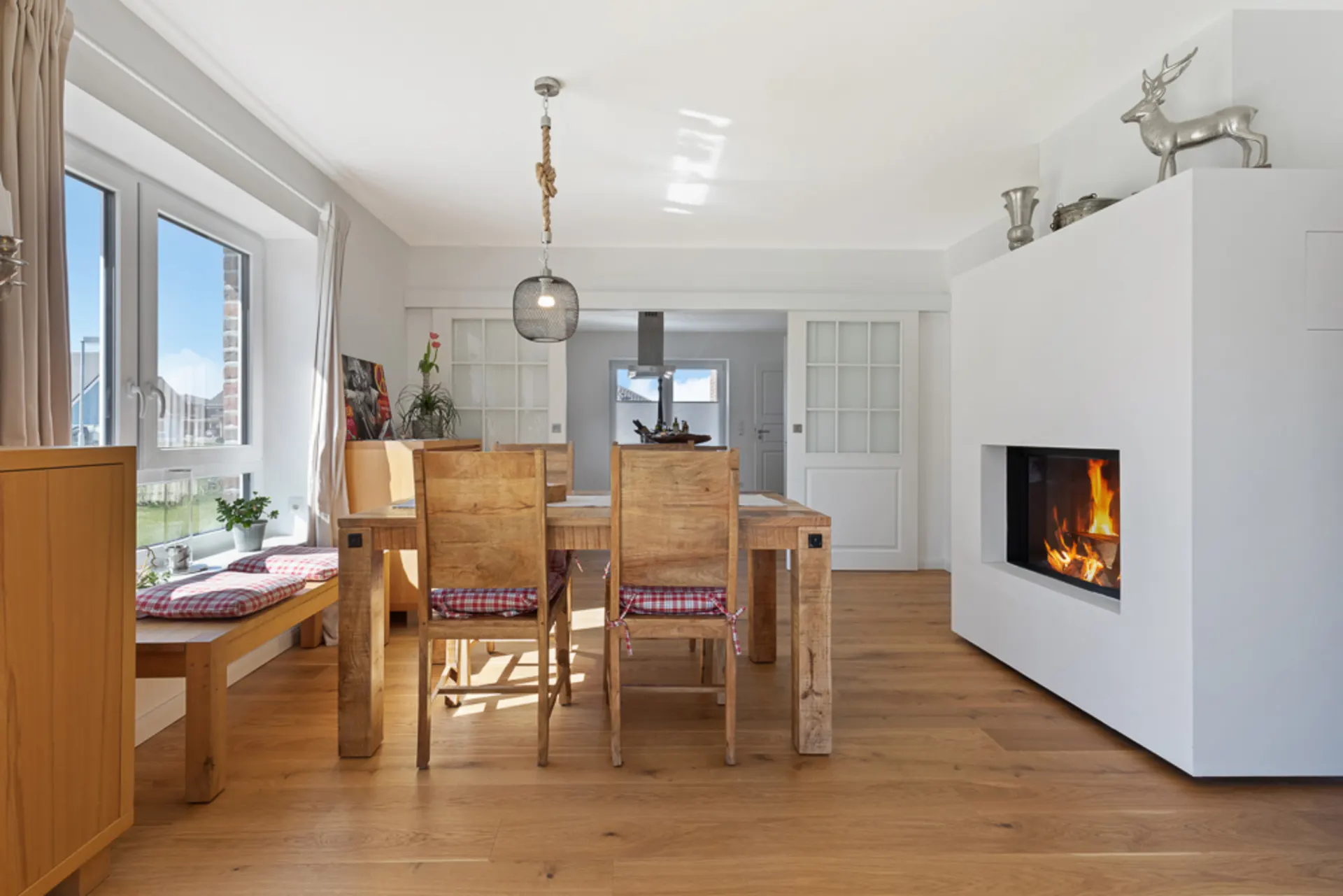 Helles Esszimmer mit Holztisch, Kaminfeuer und großen Fenstern, Sitzbank am Fenster.