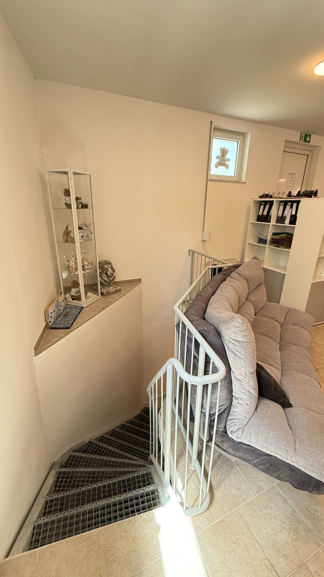 Wohnbereich mit grauem Sofa, Metalltreppe und Regal mit Dekofiguren.
