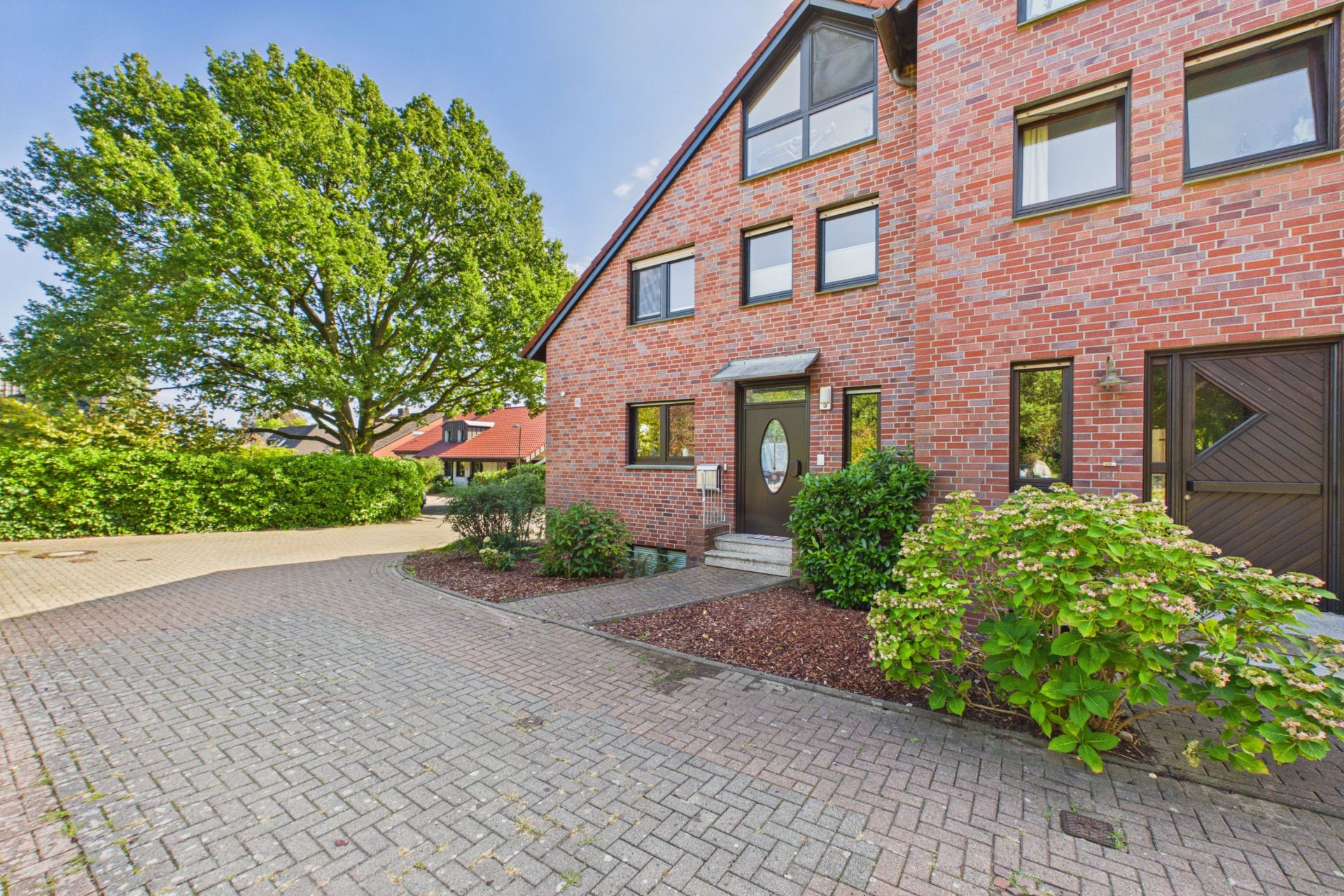 Eingang eines Backsteinhauses mit großen Fenstern, Einfahrt und grünen Büschen vor dem Haus.