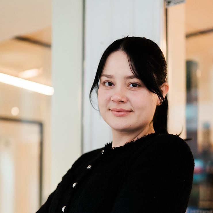 Frau mit dunklen Haaren und schwarzer Jacke steht vor einer Glastür in einem Innenraum.