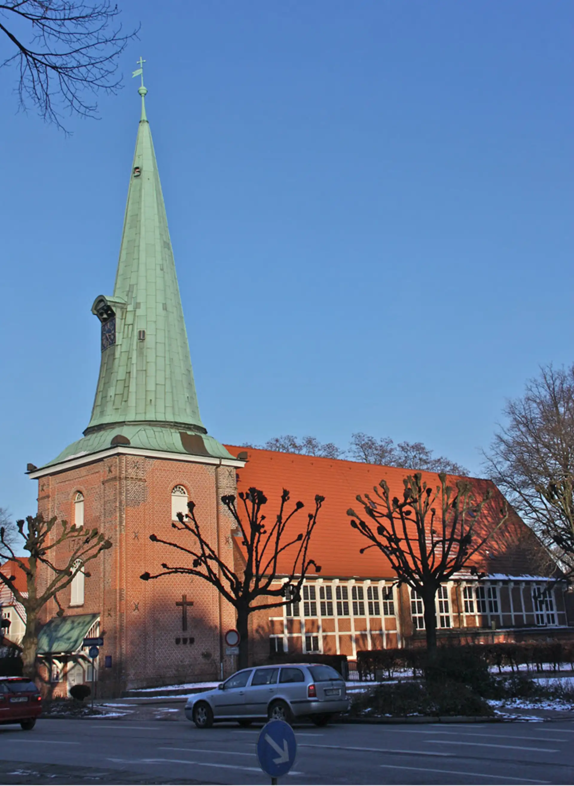 Backsteinkirche mit hohem grünen Turm und Autos davor bei klarem Himmel.