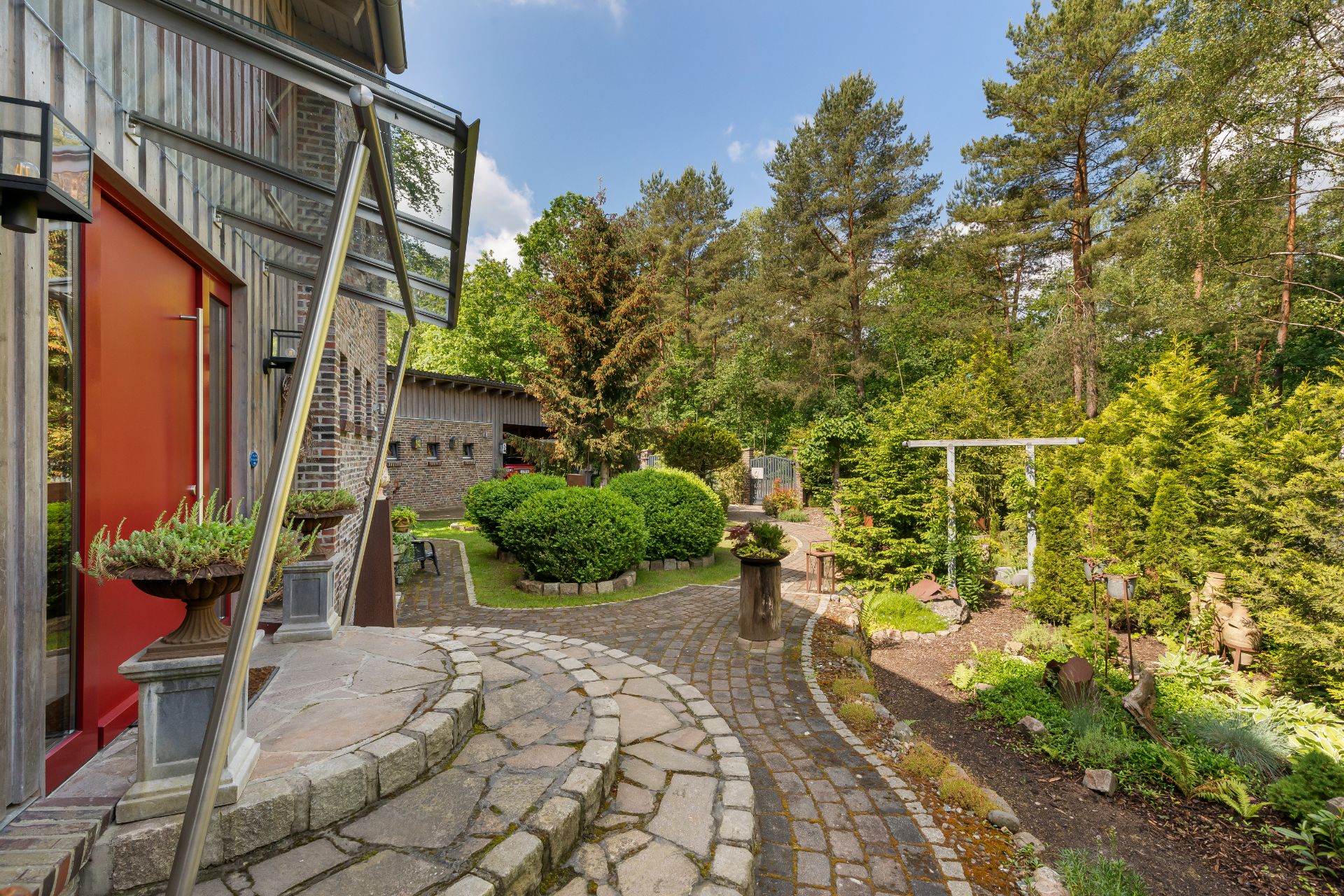 Gepflegter Garten mit Steinwegen, Pflanzen, roter Tür und Bäumen im Hintergrund.