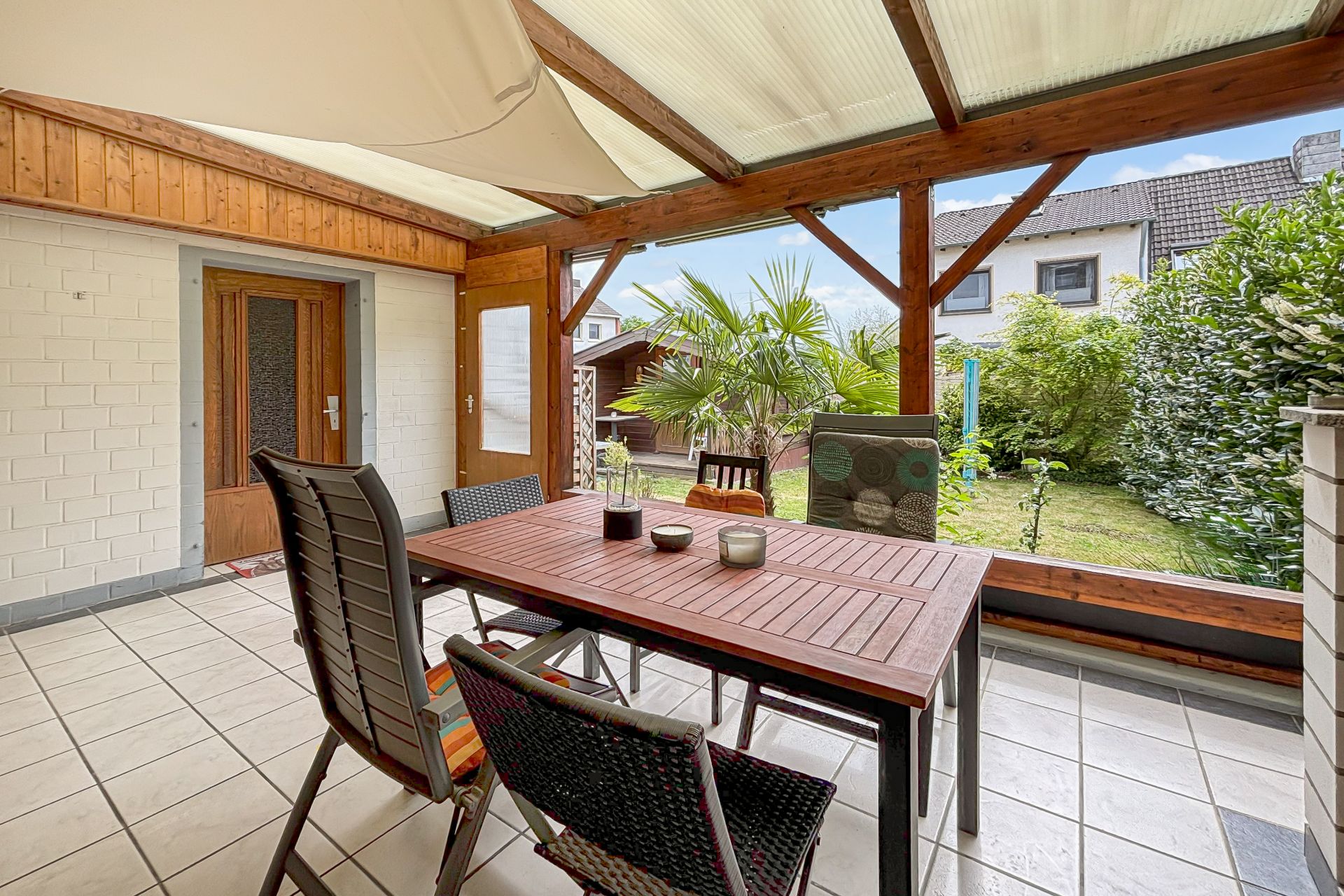 Überdachte Terrasse mit Holztisch, Stühlen und Blick auf den Garten mit Palmen.