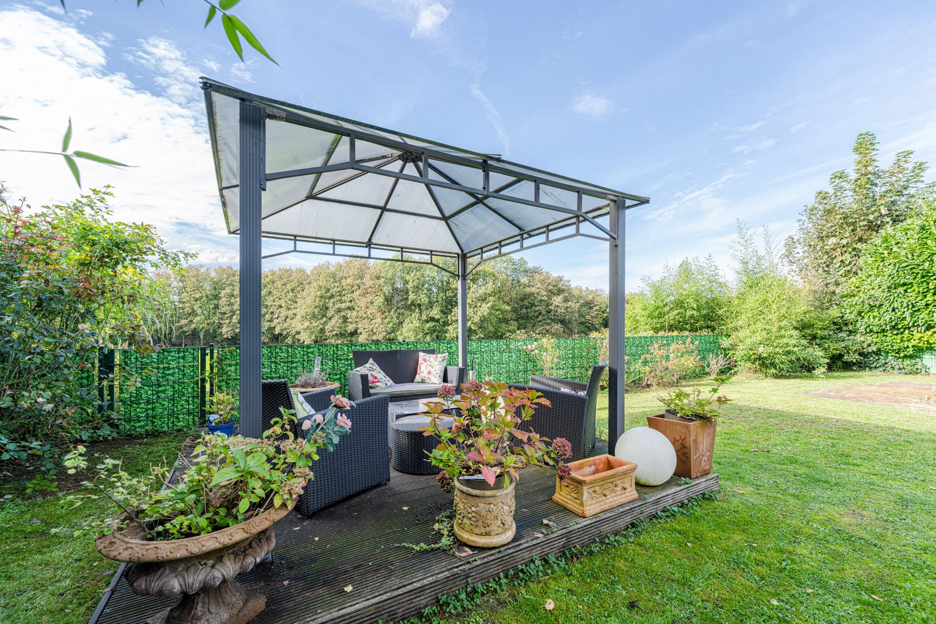 Überdachter Sitzbereich im Garten mit Gartenmöbeln und verschiedenen Pflanzgefäßen.