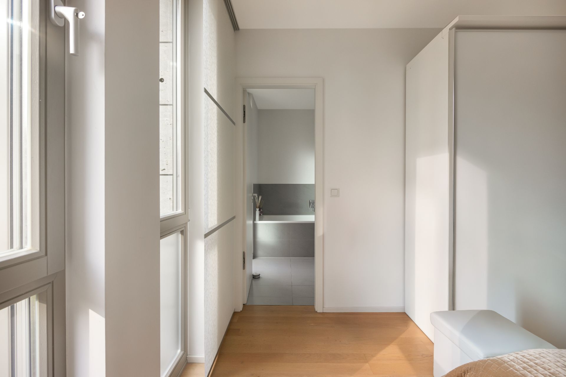 Heller Flur mit Holzboden, großen Fenstern und Blick auf ein Badezimmer mit Badewanne.