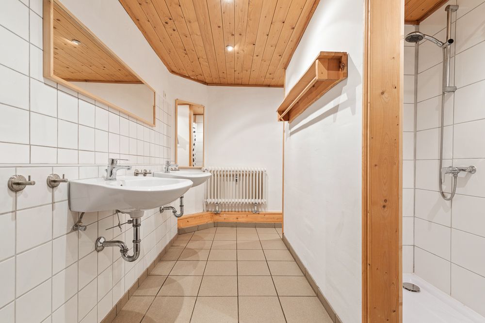 Helles Bad mit zwei Waschbecken, Dusche, Spiegeln, Holzdecke und Fliesenboden.