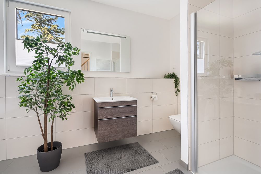 Modernes Badezimmer mit Waschbecken, Spiegel, Dusche, Topfpflanze und Fenster.