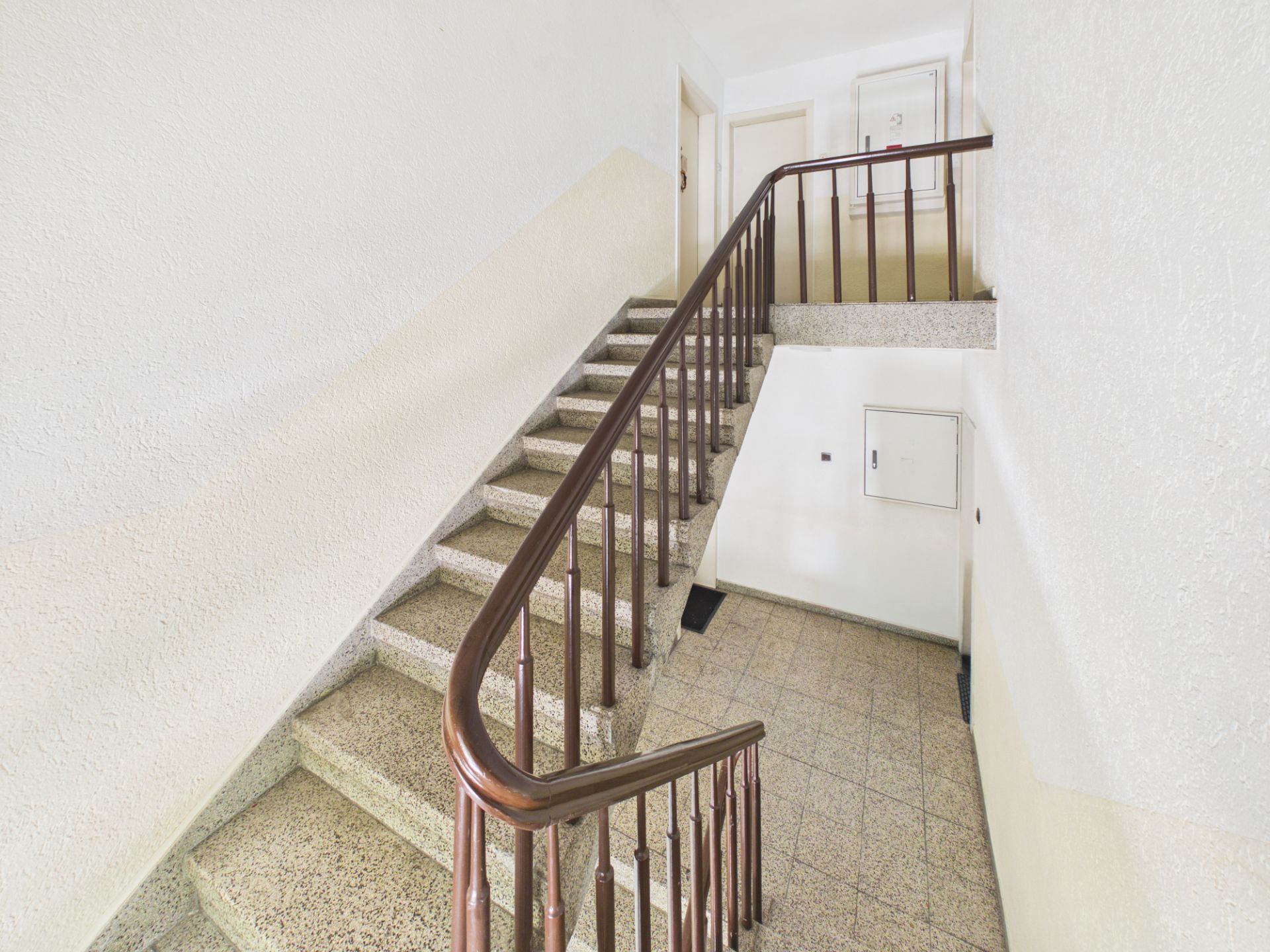 Helles Treppenhaus mit steinernen Stufen und braunem Handlauf in einem Wohngebäude.