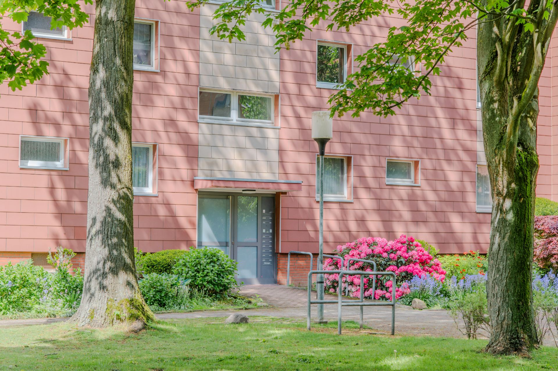 Eingang eines Wohnhauses mit roten Fassaden, Blumenbeeten und Bäumen im Vordergrund.