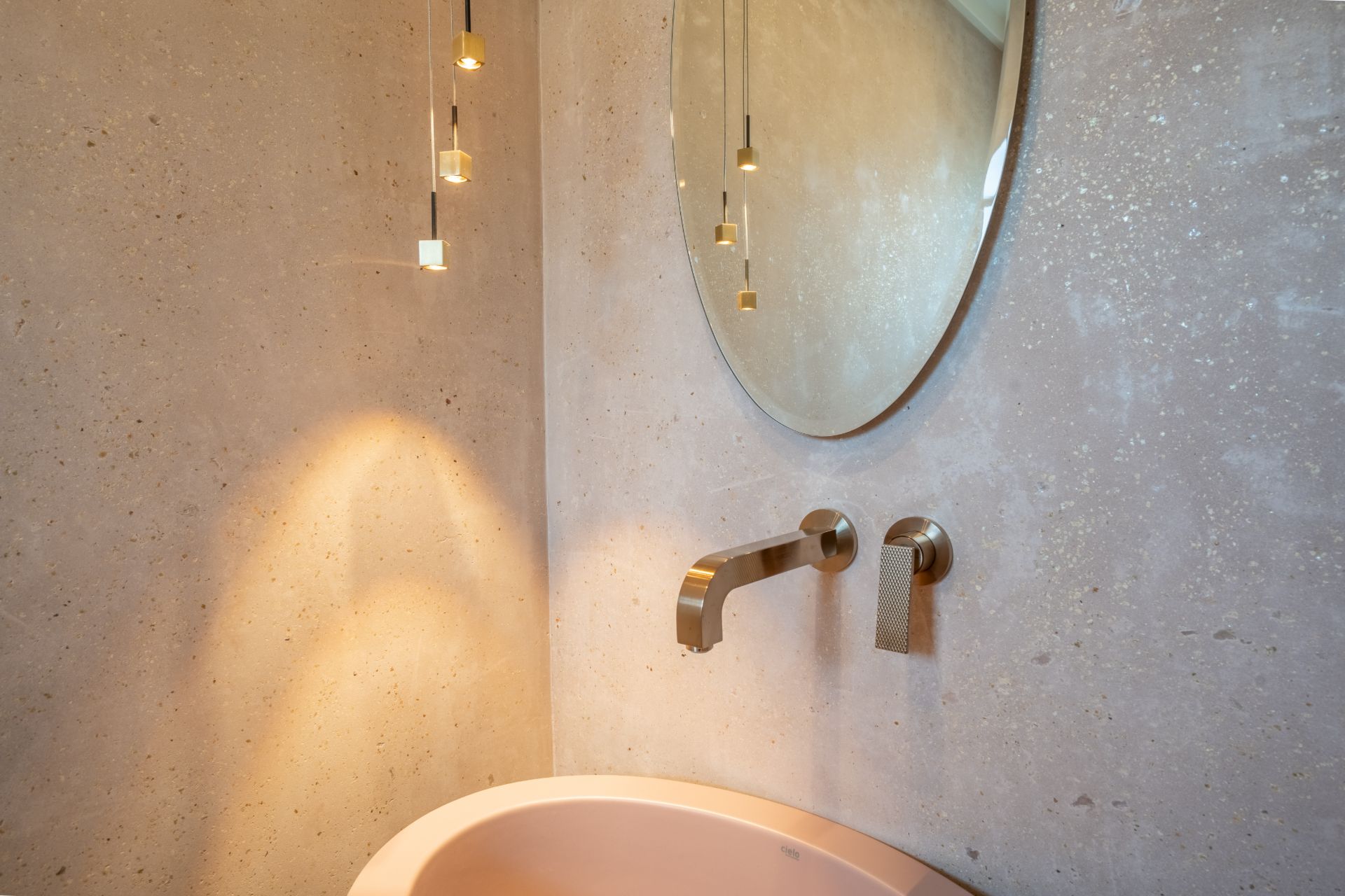 Waschbecken mit Wandarmatur, rundem Spiegel und Hängelampen an einer hellen Wand.