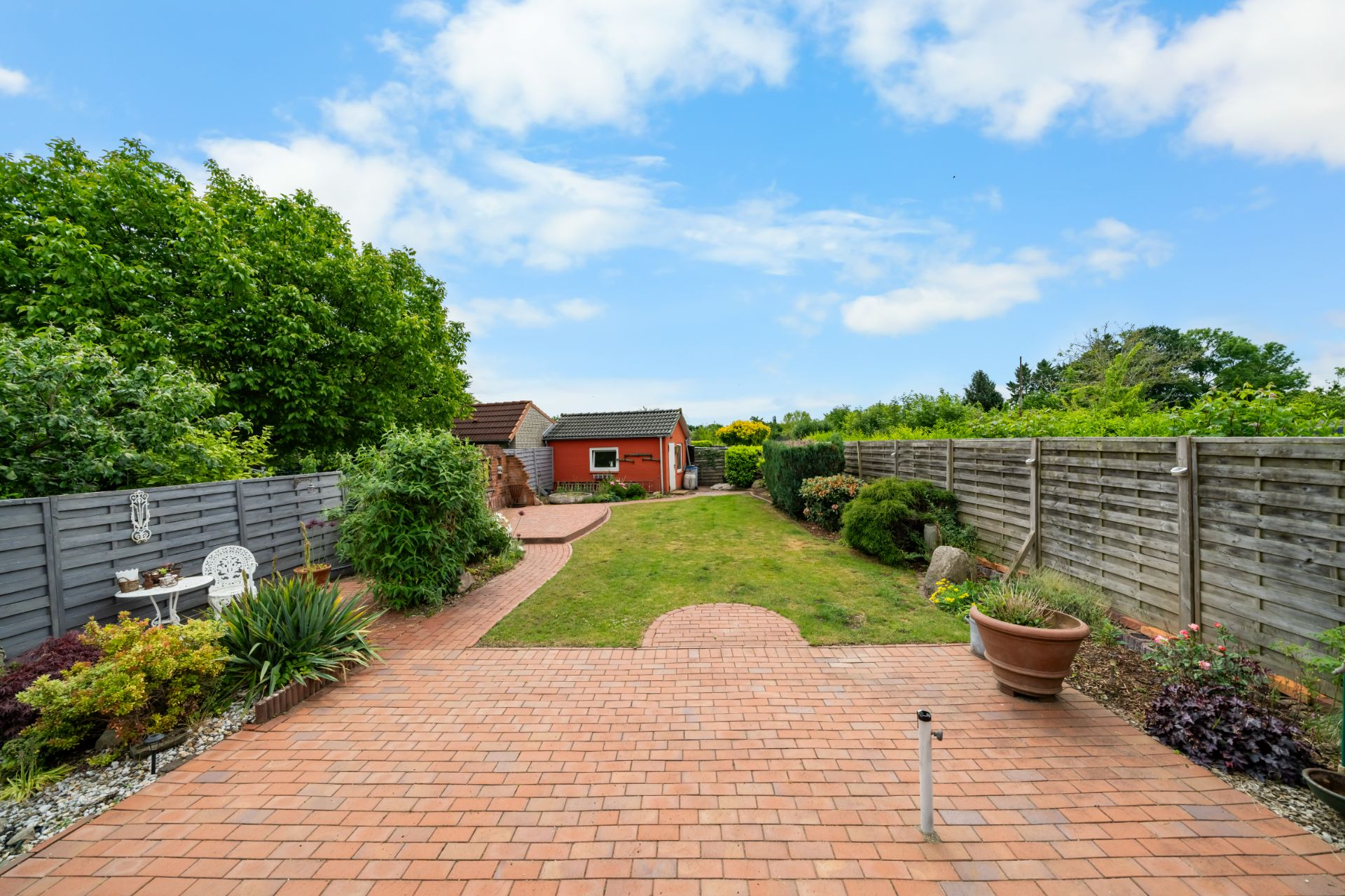 Gepflasterte Terrasse mit Garten, Pflanzen, Zäunen und Gartenhaus im Hintergrund.