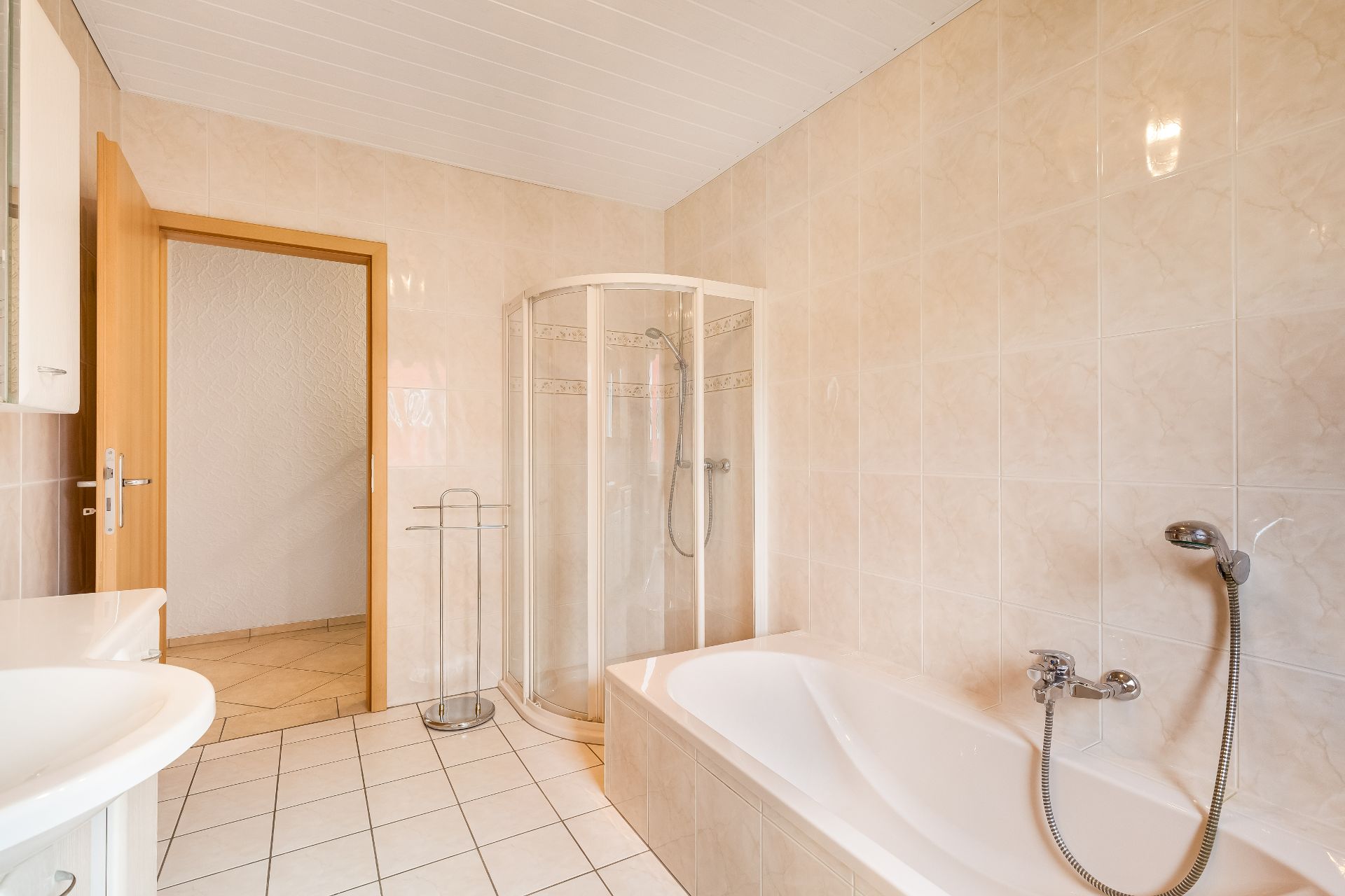 Helles Badezimmer mit Badewanne, Dusche und Waschbecken, gefliest und mit offenstehender Holztür.