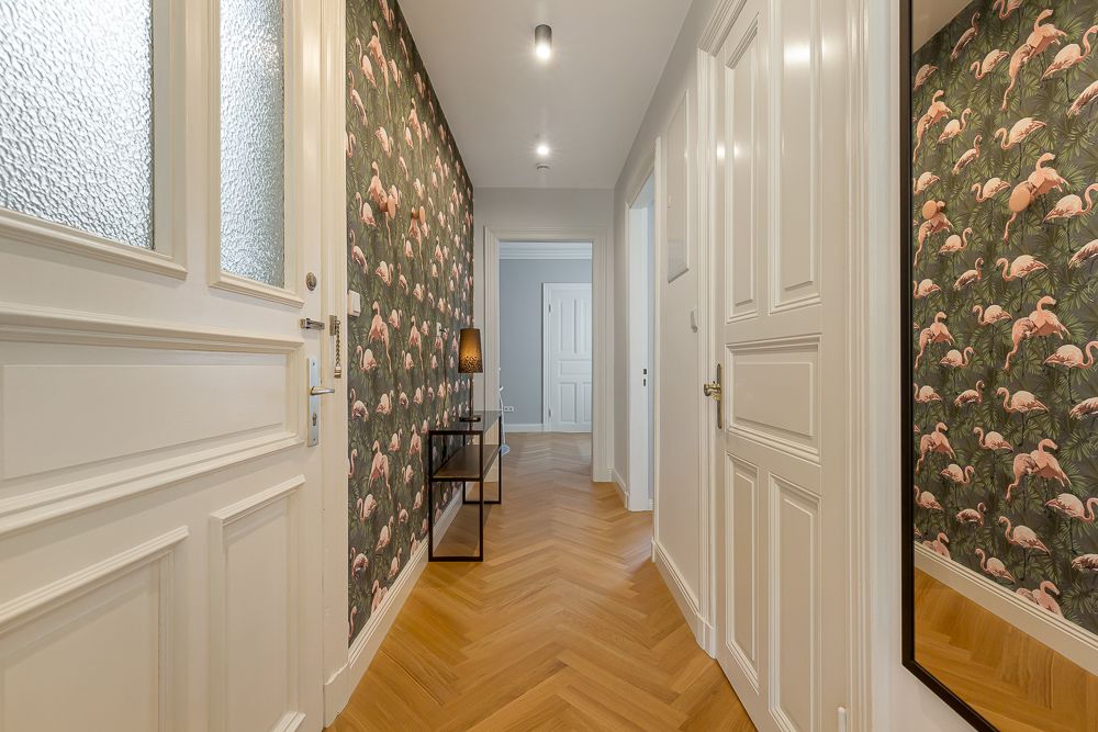 Heller Flur mit Flamingo-Tapete, Holzboden, Spiegel und kleiner Konsole mit Lampe.