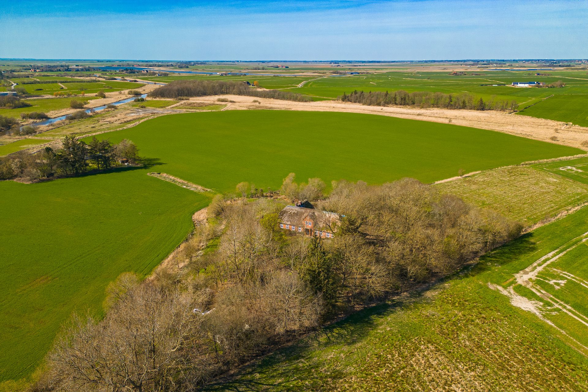 Backsteinhaus mit Reetdach, umgeben von Bäumen und weiten grünen Feldern, aus der Vogelperspektive.
