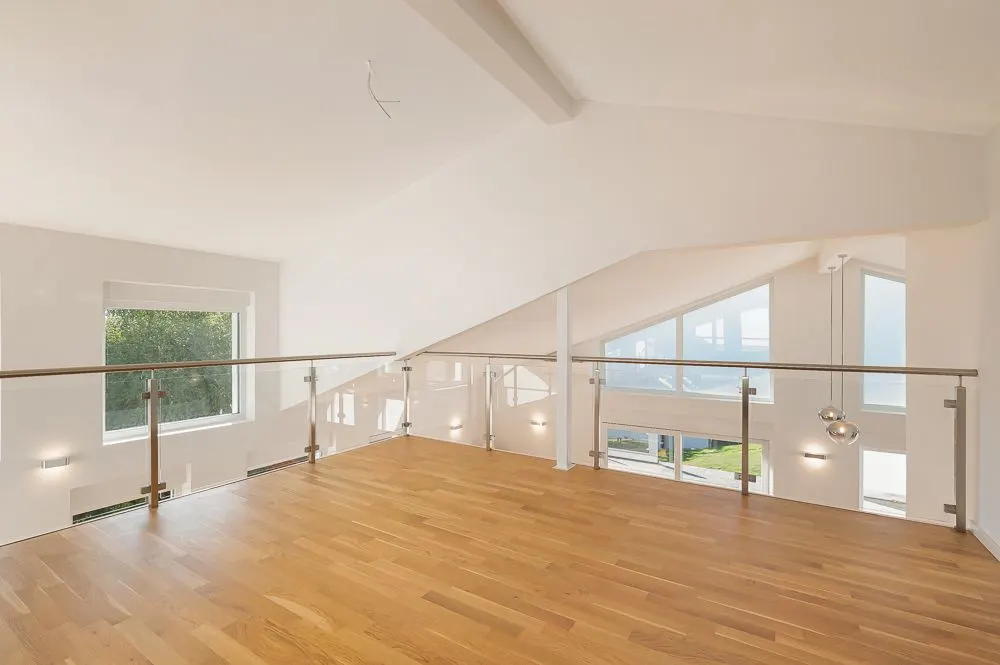 Leerer Raum mit Holzfußboden und Glasgeländer, große Fenster, viel Tageslicht.