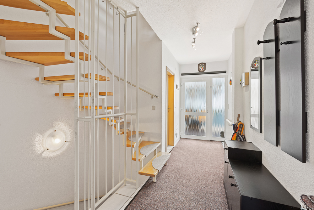 Heller Flur mit Holztreppe, weißen Wänden und Garderobe neben der Eingangstür.