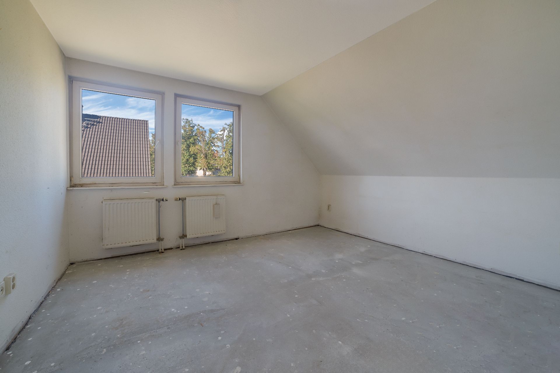 Leeres Zimmer mit zwei Fenstern, weiß gestrichenen Wänden und blankem Boden.