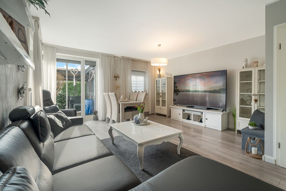 Helles Wohnzimmer mit schwarzem Sofa, Couchtisch, großem Fernseher und Esstisch vor Terrassentür.