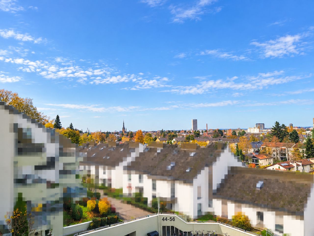 Blick auf weiße Reihenhäuser und herbstliche Bäume unter blauem Himmel.