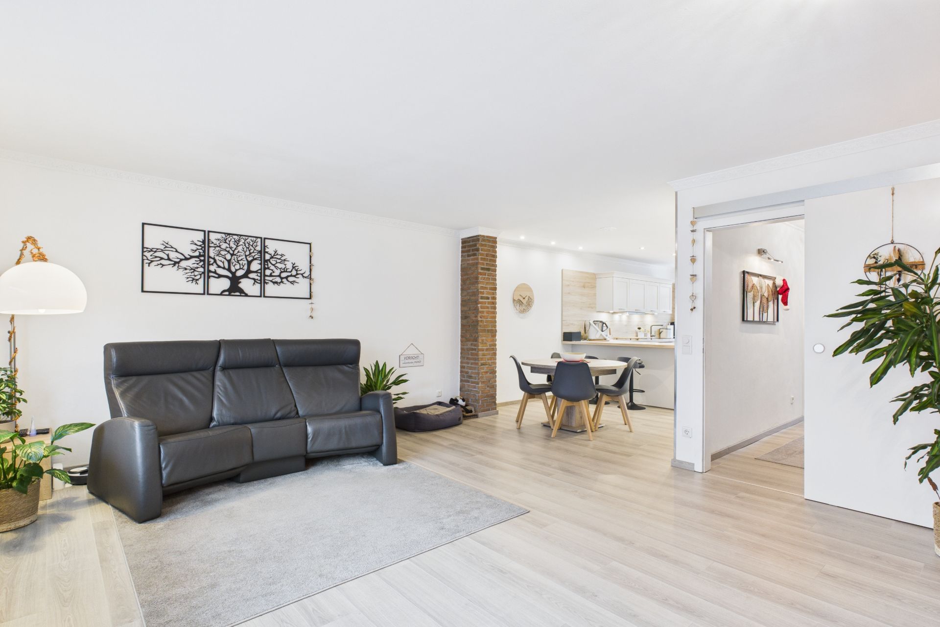 Modernes Wohnzimmer mit schwarzem Sofa, Esstisch, Pflanzen und offener Küche auf hellem Holzboden.