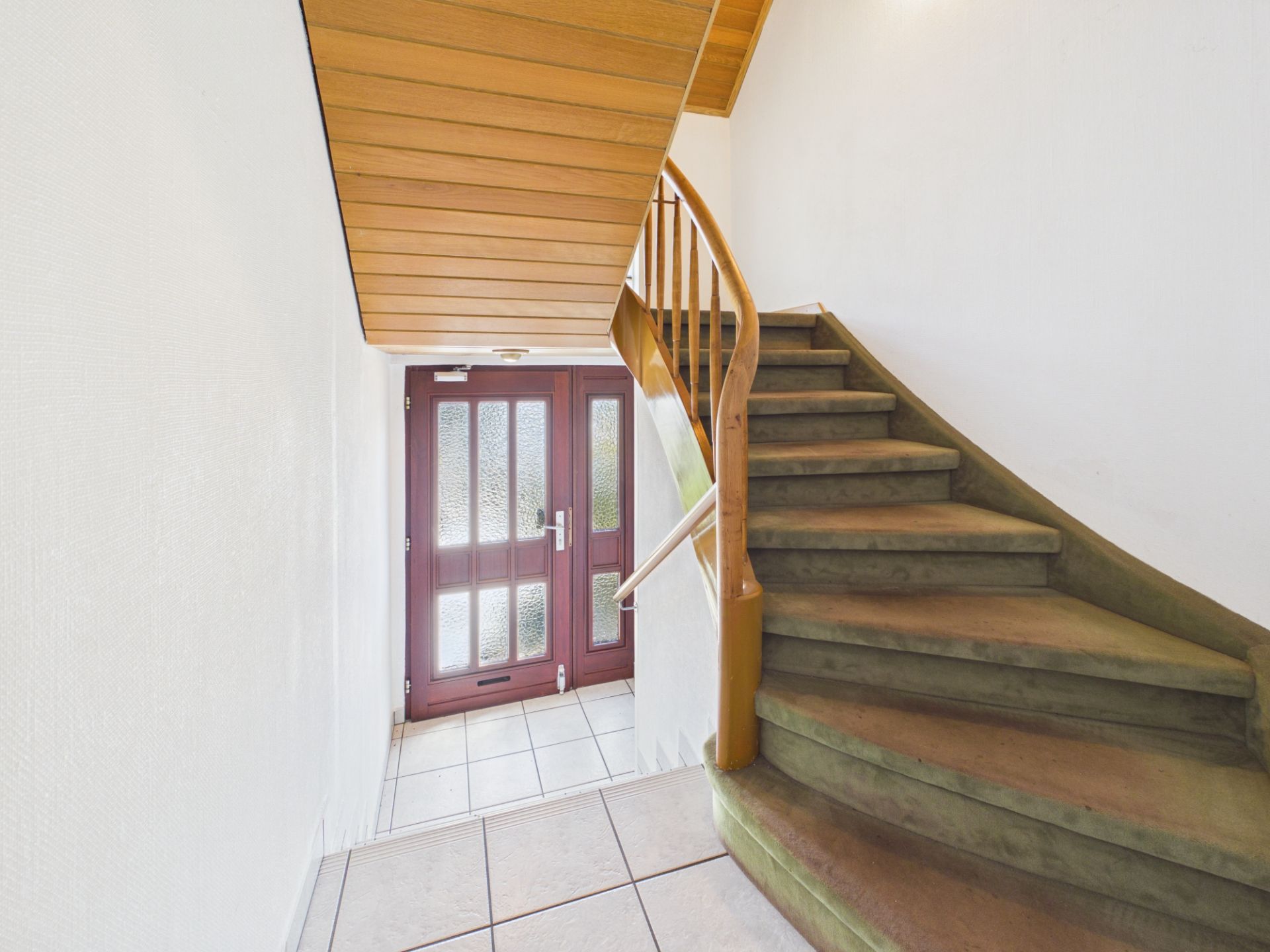 Treppe mit grünem Teppichbelag, daneben eine Glastür mit Holzrahmen in einem Flur.