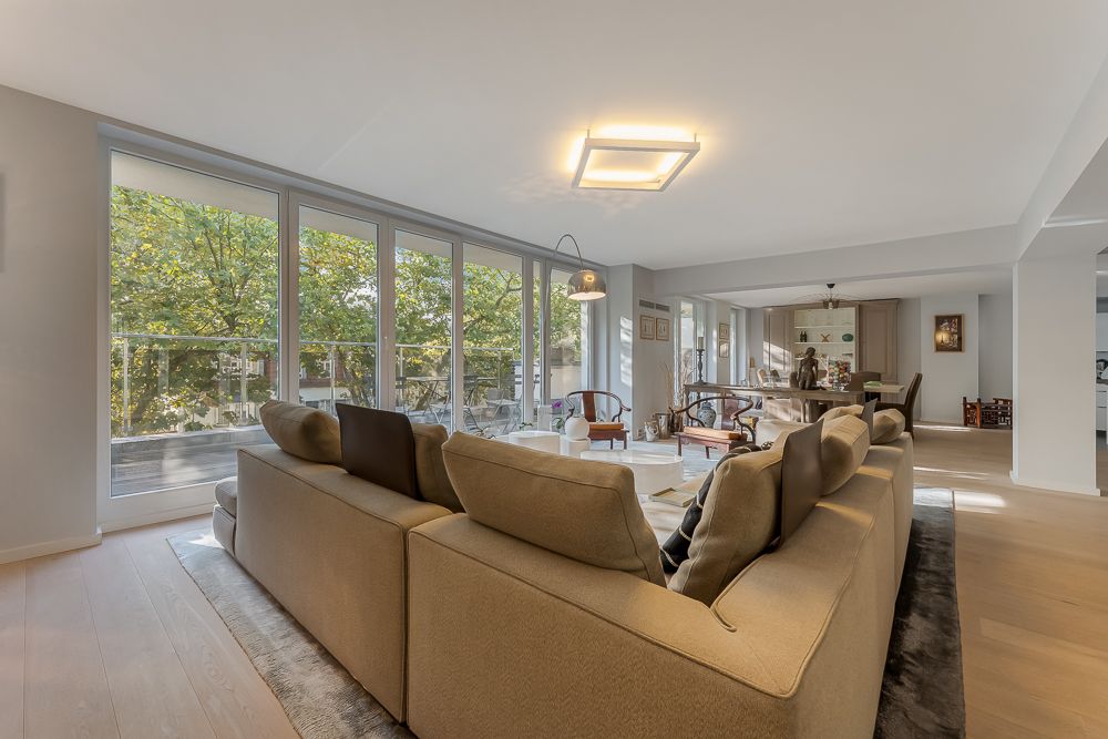 Helles Wohnzimmer mit großer Couch und bodentiefen Fenstern zu einem Balkon mit Blick auf Bäume.