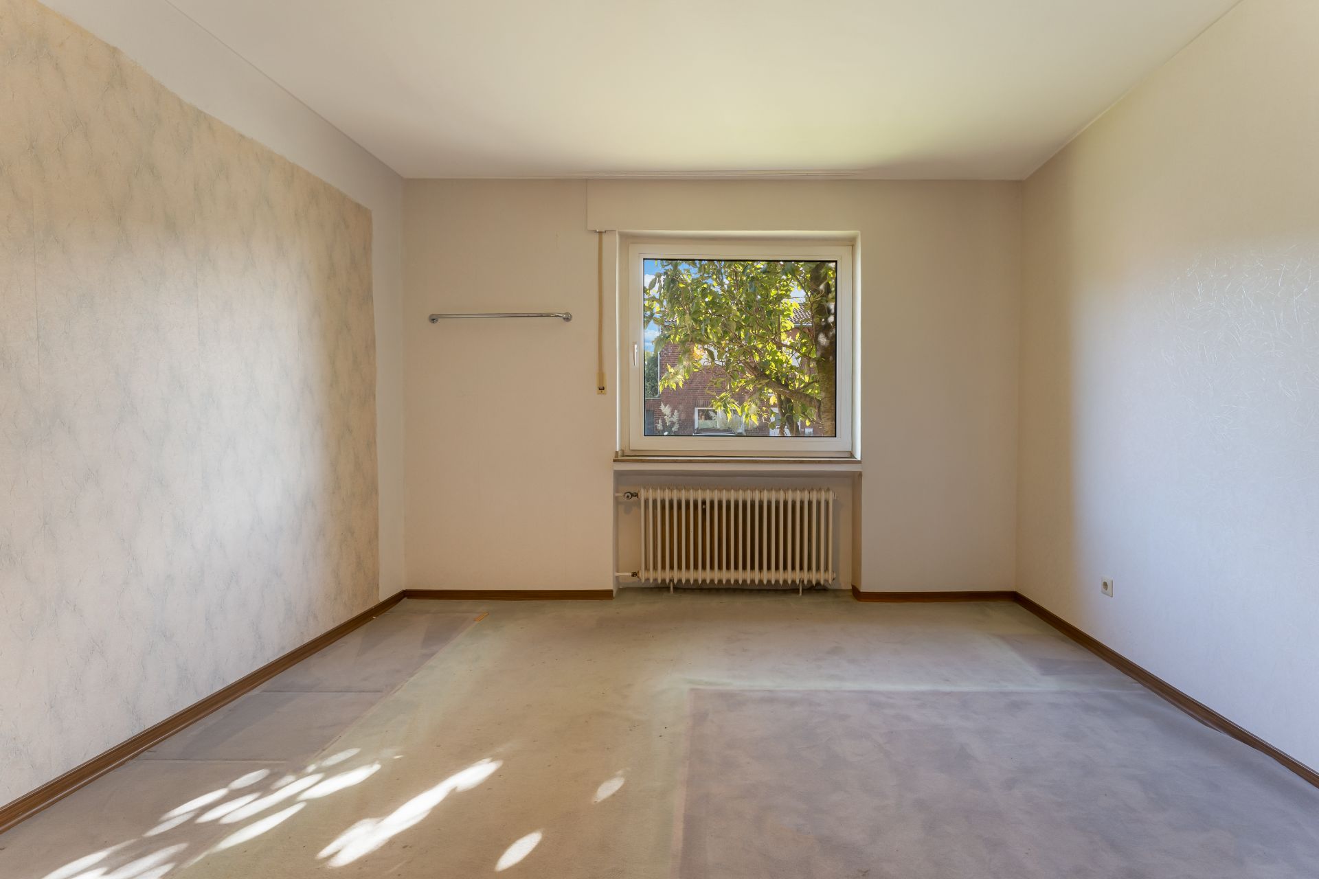 Leeres Zimmer mit Teppichboden, Fenster und Heizung unter dem Fenster.