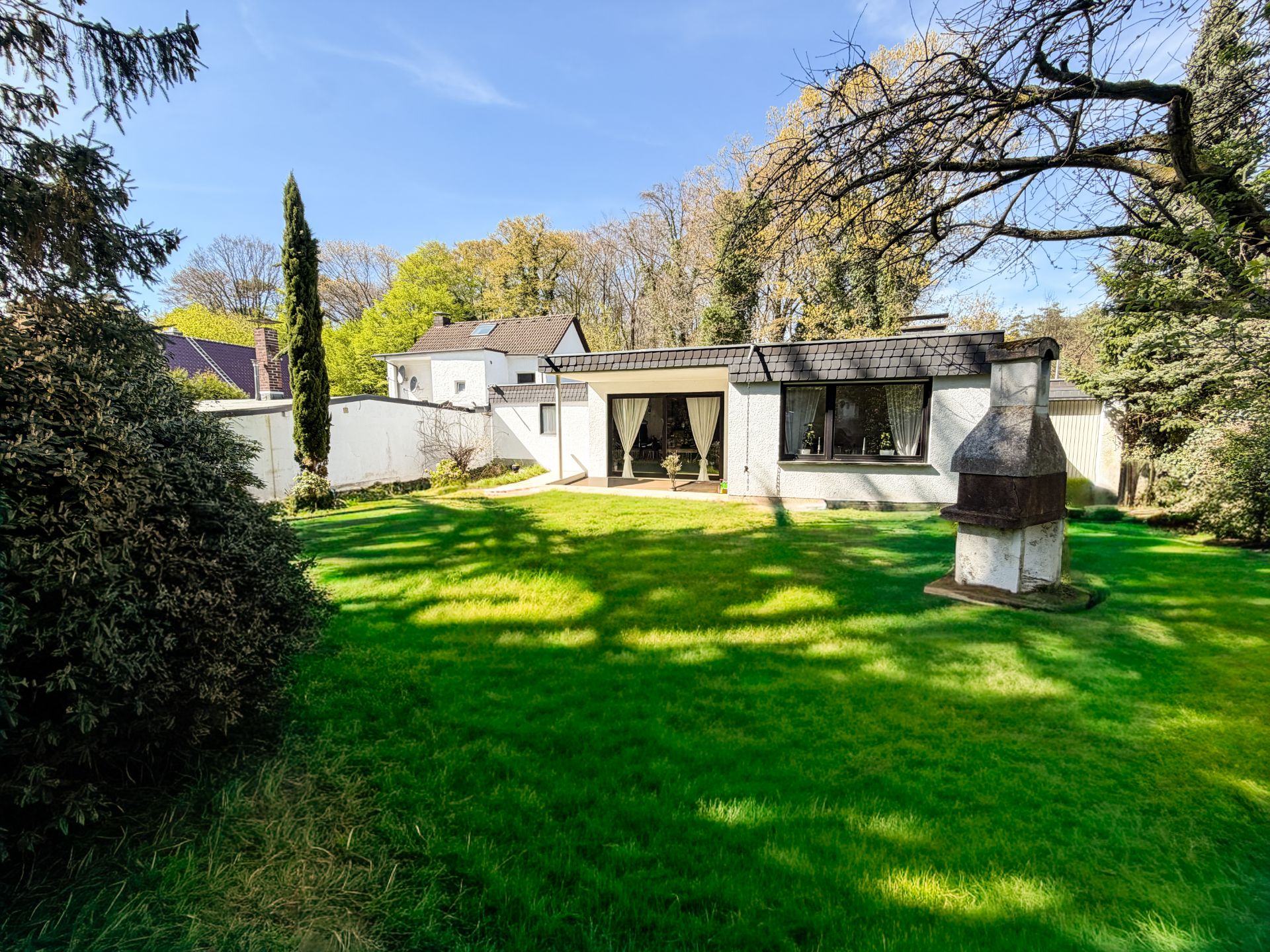 Grüner Garten mit Rasen, Bäumen, einem Grillkamin und einem weißen Flachbauhaus im Hintergrund.