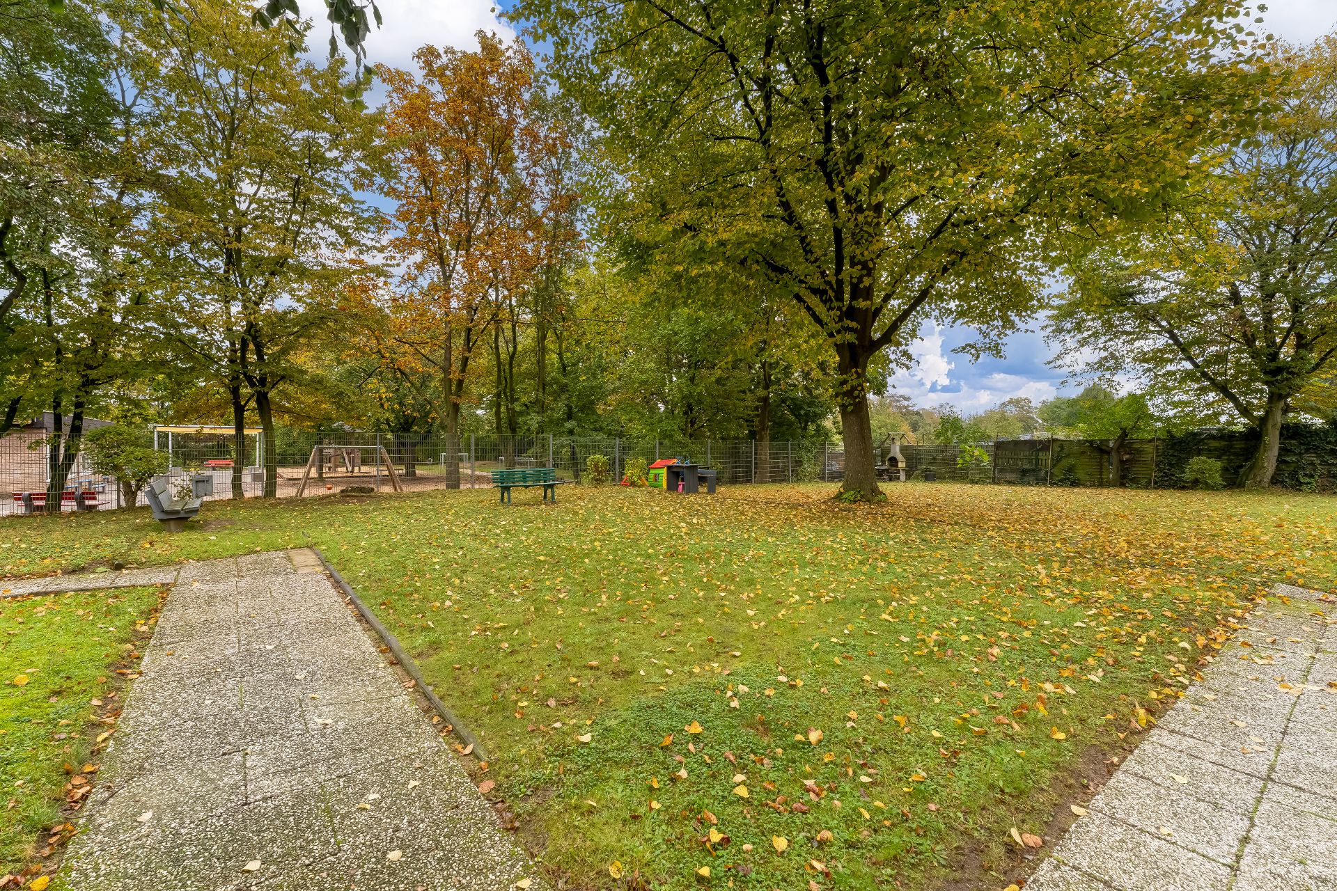 Grüne Rasenfläche mit Wegen, Bäumen, Bänken und einem Spielplatz im Hintergrund.