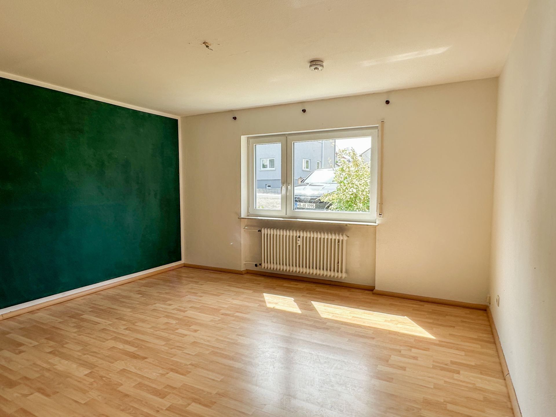 Helles, leeres Zimmer mit Laminatboden, grün gestrichener Wand und großem Fenster mit Heizkörper.