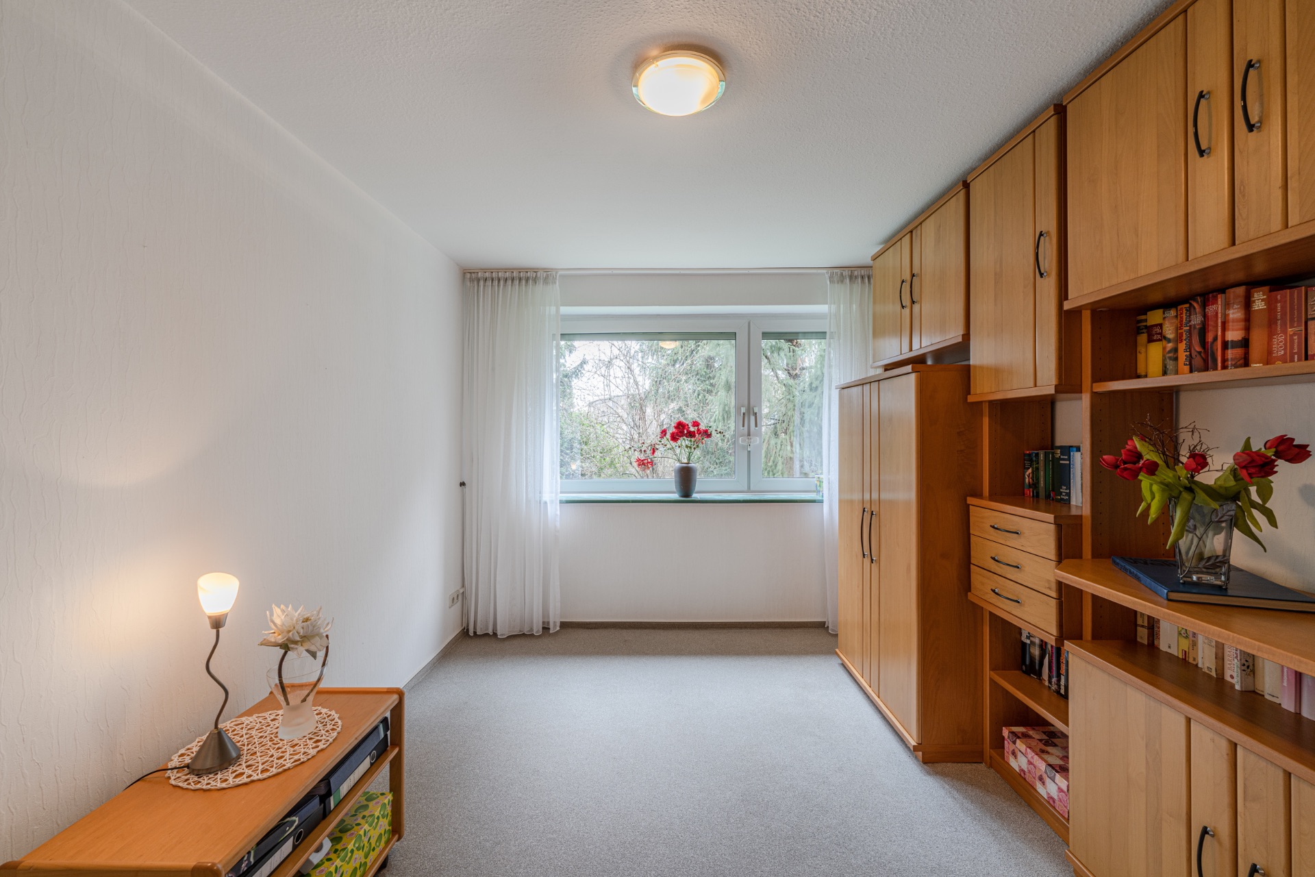 Helles Zimmer mit Teppichboden, Holzregalen, Fenster und Blumenvasen auf Möbeln.