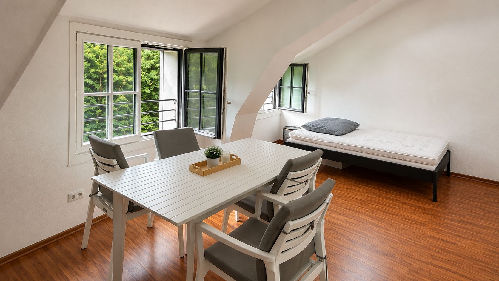Helles Zimmer mit Holzboden, Esstisch, vier Stühlen, Bett und geöffnetem Fenster.