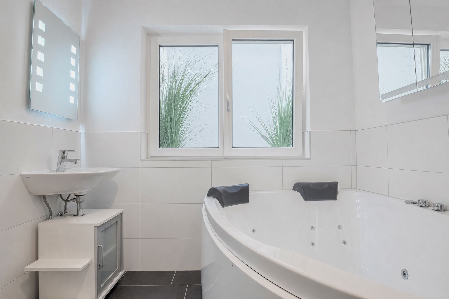 Modernes Badezimmer mit großer weißen Eckbadewanne, Waschbecken und Fenster mit Pflanzen draußen.