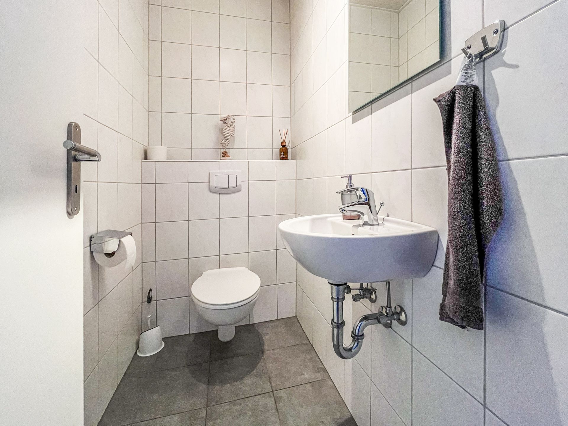 Kleines, weiß gefliestes Badezimmer mit Wand-WC, Waschbecken, Spiegel und Handtuch.