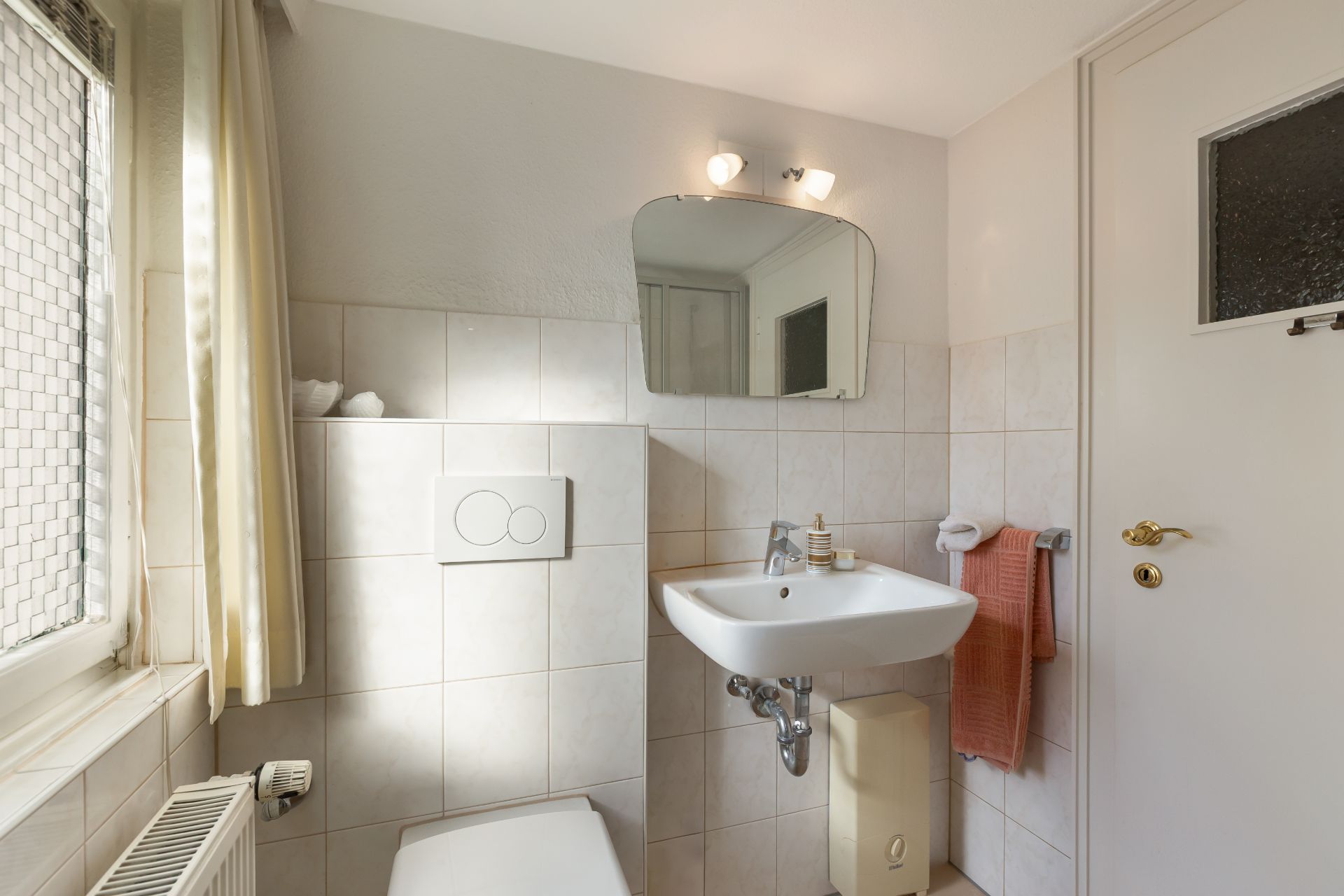 Helles Badezimmer mit Wandtoilette, Waschbecken, Spiegel und Handtuch neben der Tür.
