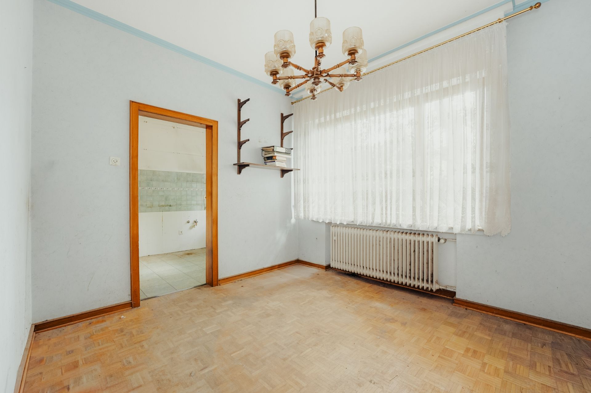Leeres Zimmer mit Holzfußboden, großem Fenster, Heizkörper, Regal und Kronleuchter.