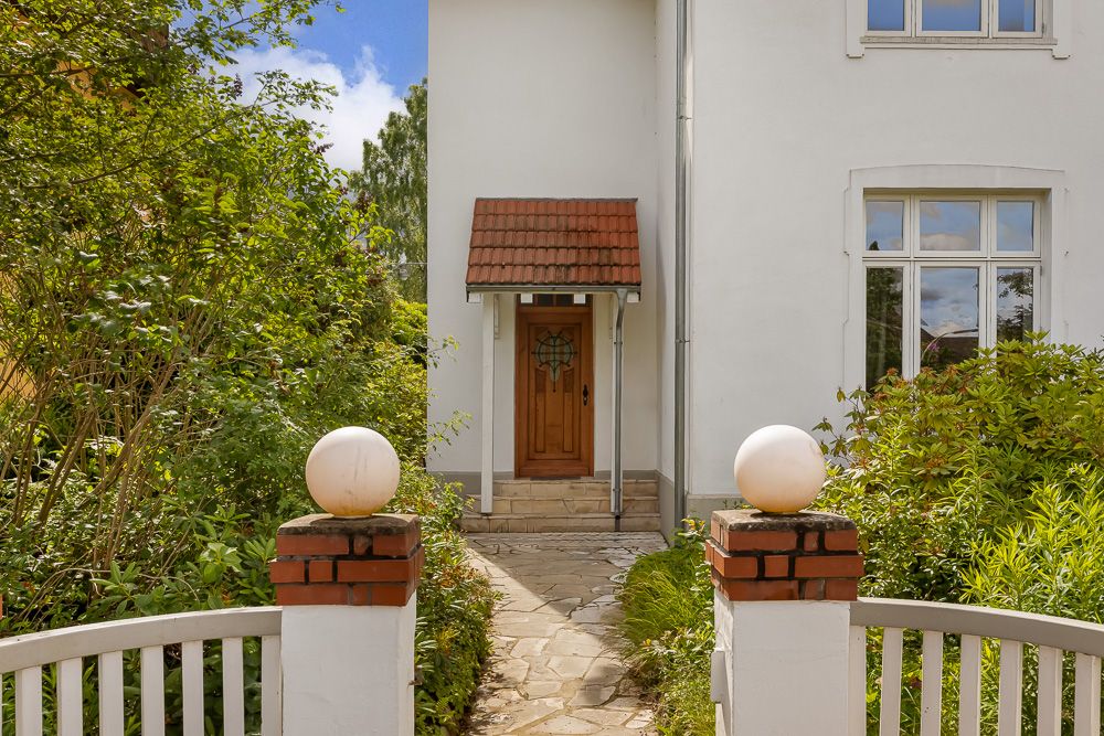 Eingang eines weißen Hauses mit Holztür, Gartenweg und Bepflanzung.