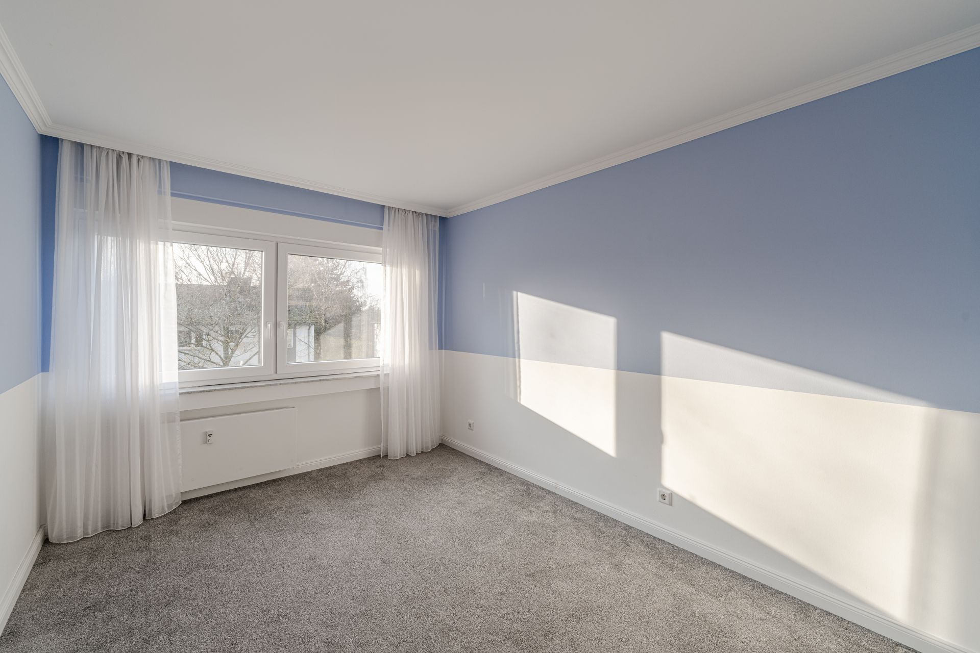 Leeres Zimmer mit blau-weißen Wänden, Teppichboden und großem Fenster mit weißen Vorhängen.