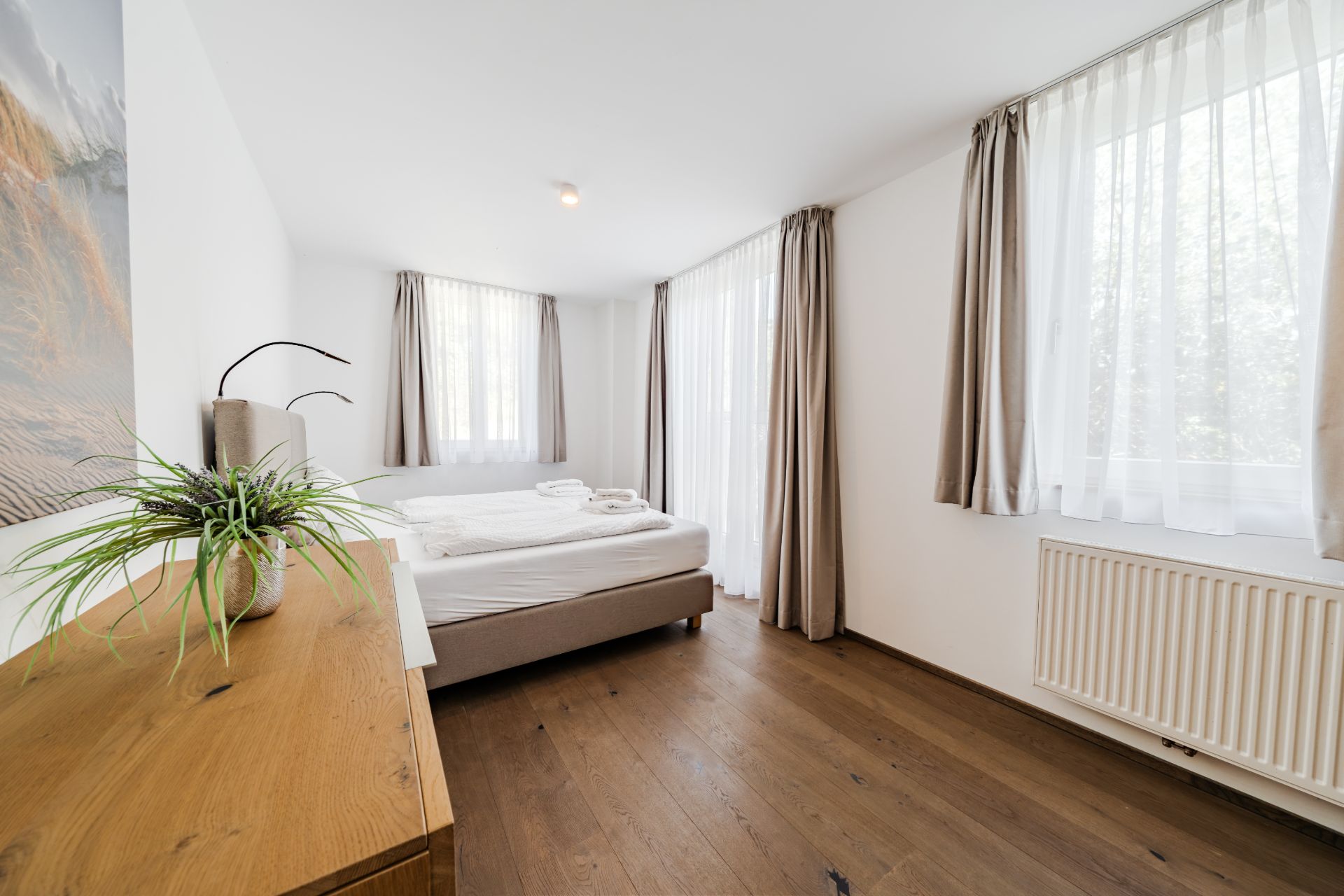 Helles Schlafzimmer mit Doppelbett, Holzboden, Kommode und drei Fenstern mit Vorhängen.