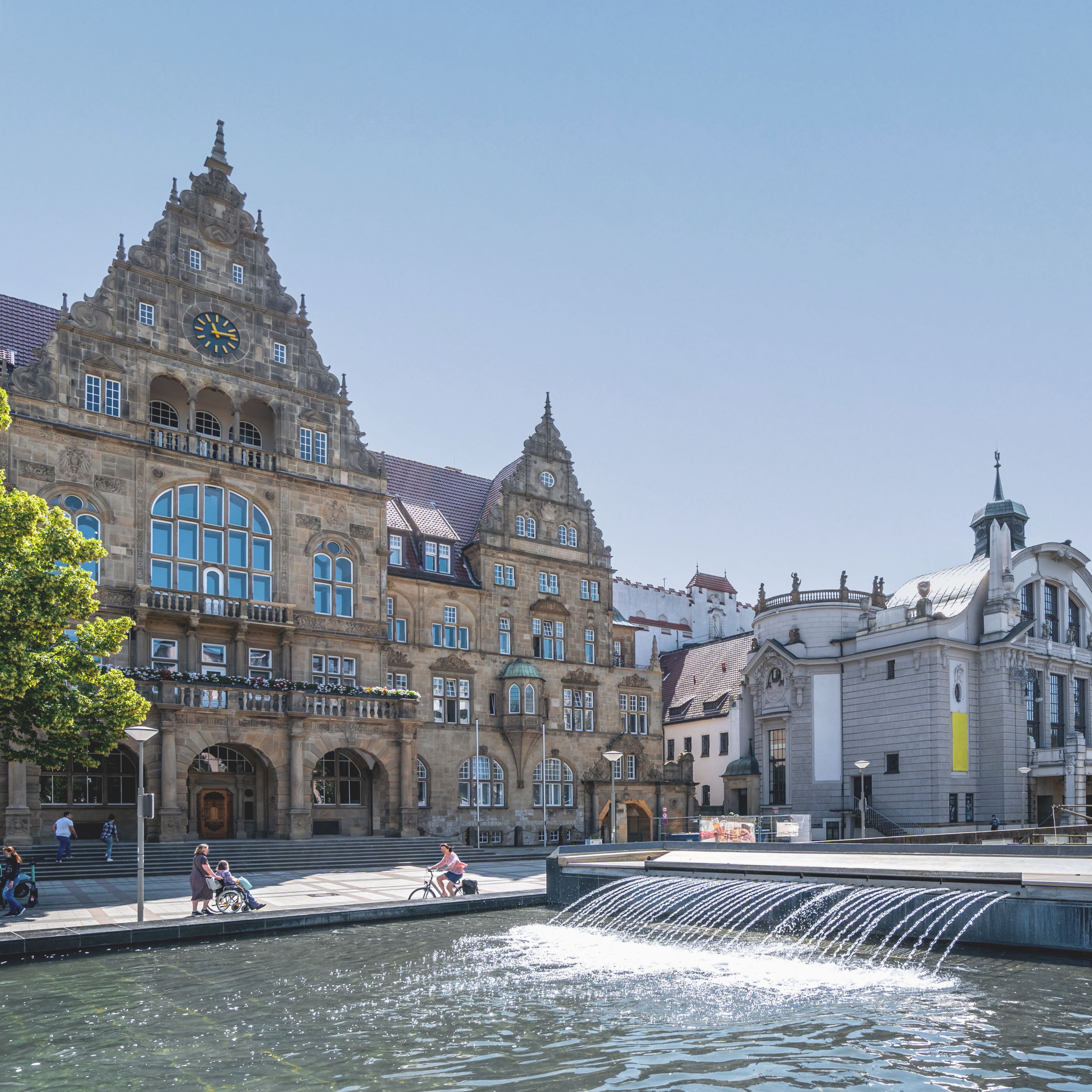 Historisches Gebäude mit Uhrturm, Wasserbecken und Springbrunnen im Vordergrund bei sonnigem Wetter.