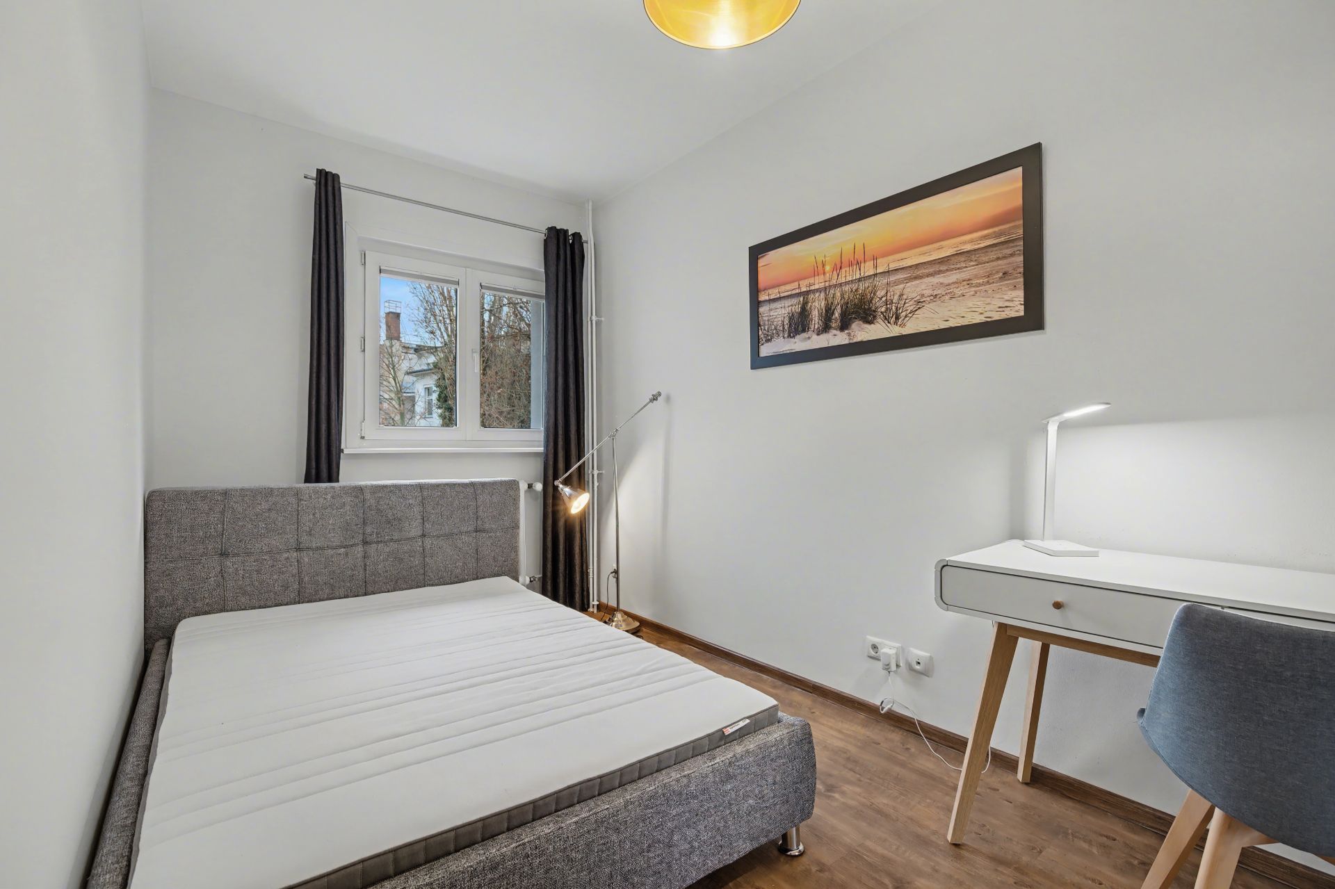 Kleines Schlafzimmer mit grauem Bett, Schreibtisch, Stuhl und Fenster mit Vorhängen.