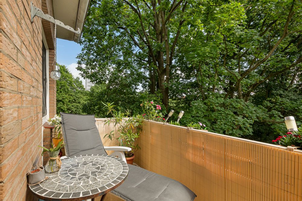 Kleiner Balkon mit Liegestuhl, rundem Tisch und Blick auf grüne Bäume.