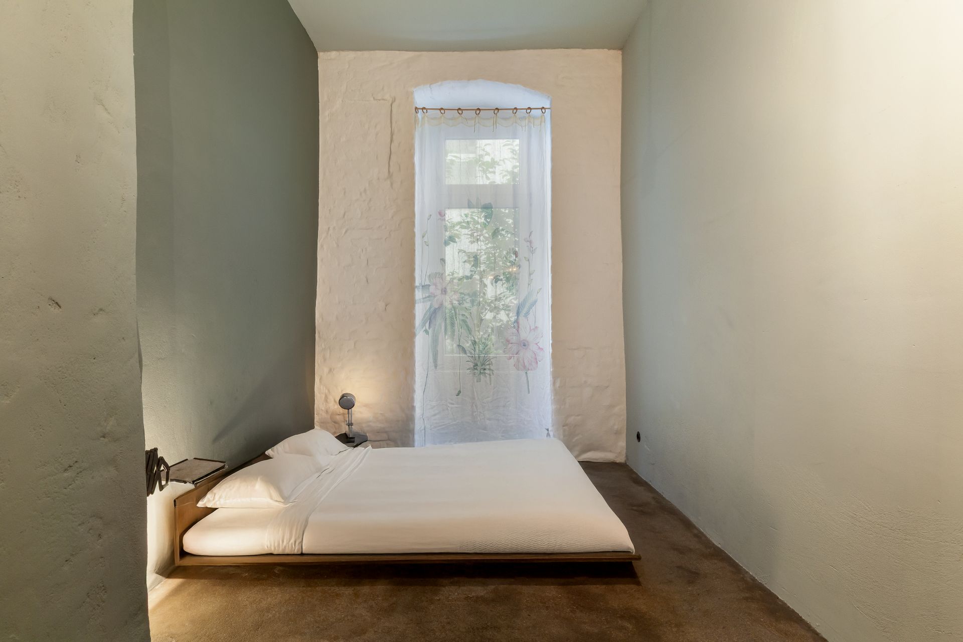Schlafzimmer mit niedrigem Bett, Nachttischlampe und Fenster mit Vorhang.