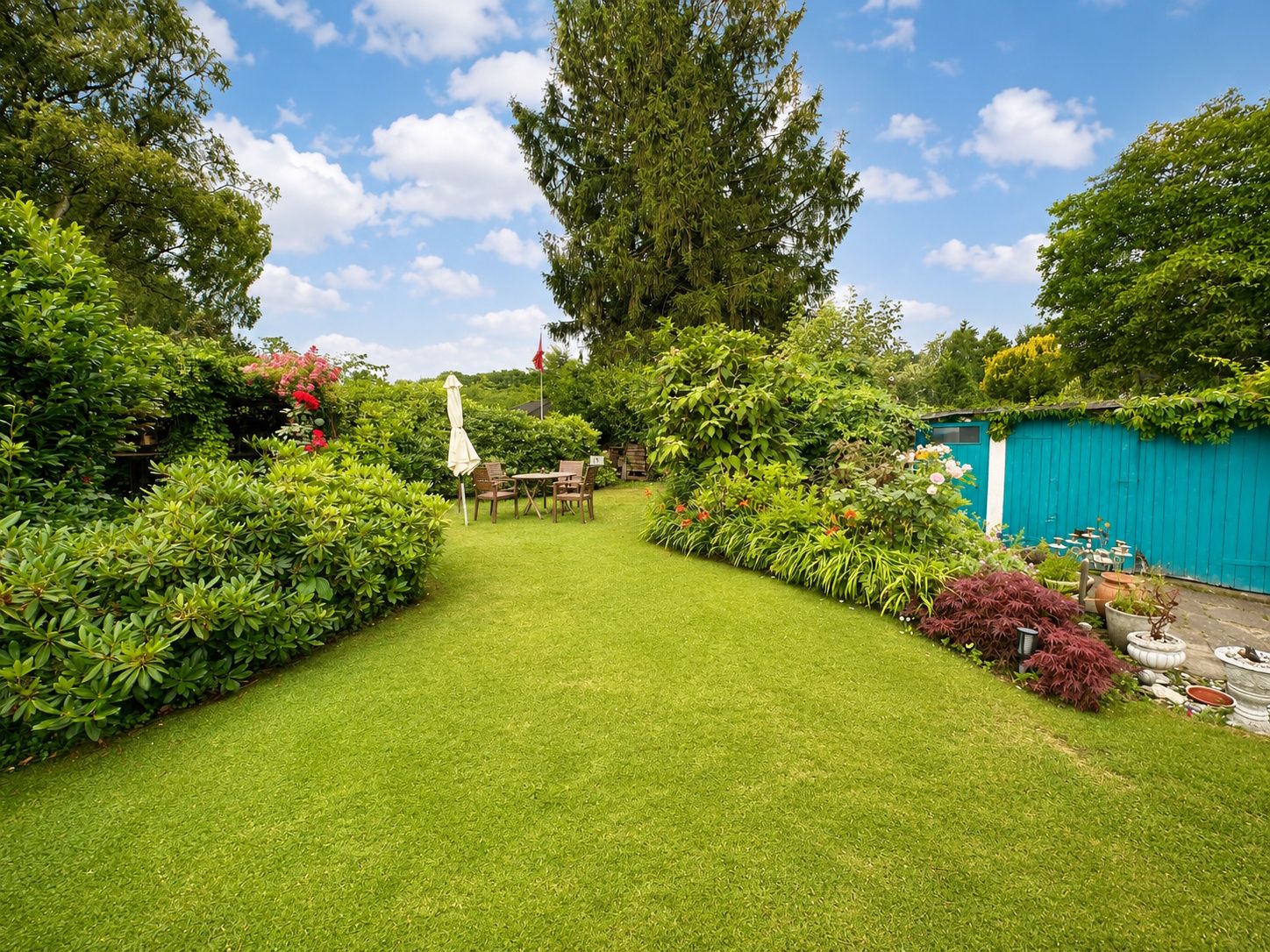 Gepflegter Garten mit Rasen, Gartenmöbeln und üppigen Sträuchern vor einem großen Baum.