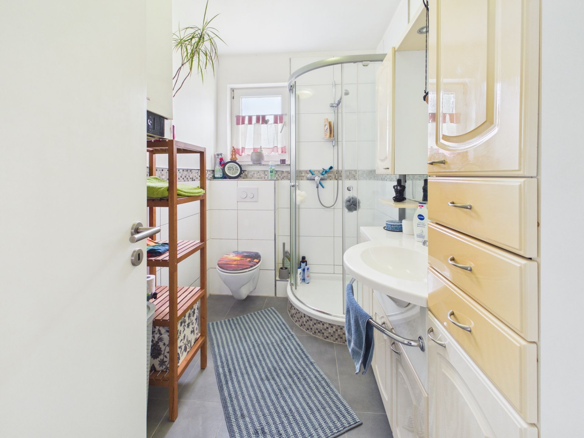 Helles Badezimmer mit Dusche, Waschbecken, WC, Regal und Schränken.