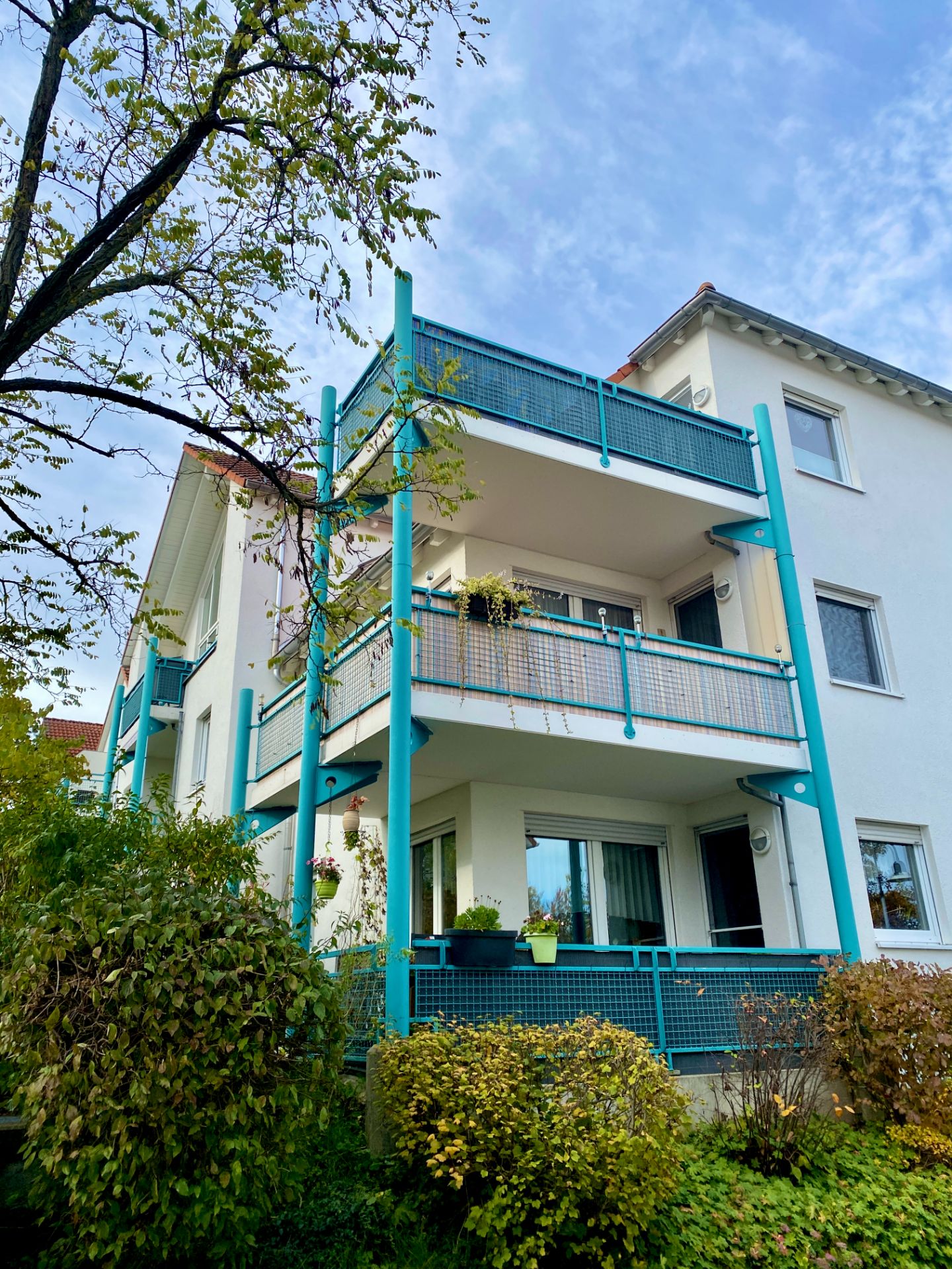 Mehrfamilienhaus mit hellen Fassaden, blauen Balkonen und Pflanzen vor dem Gebäude.