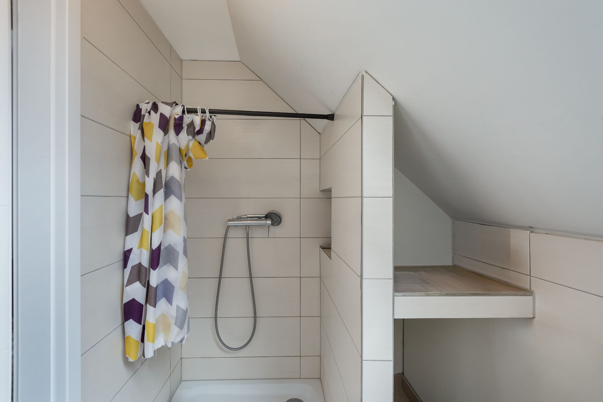 Kleine Dusche mit gemustertem Duschvorhang und gefliestem Regal unter einer Dachschräge.
