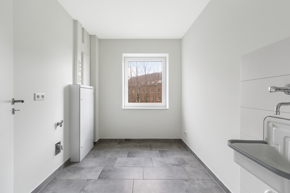 Heller leerer Raum mit Fliesenboden, Waschbecken und Fenster mit Blick auf ein Gebäude.