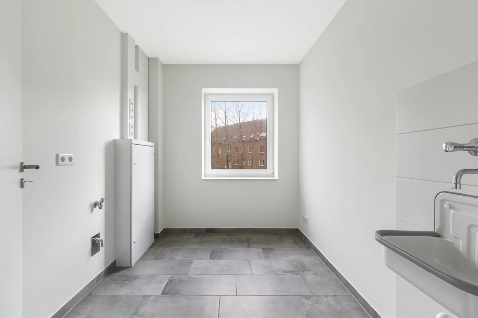Heller leerer Raum mit Fliesenboden, Waschbecken und Fenster mit Blick auf ein Gebäude.