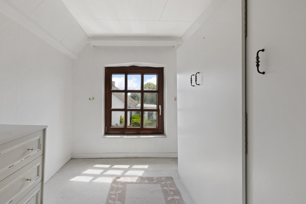 Helles Zimmer mit weißem Teppich, Fenster mit Holzrahmen, Schrankwand und Kommode.